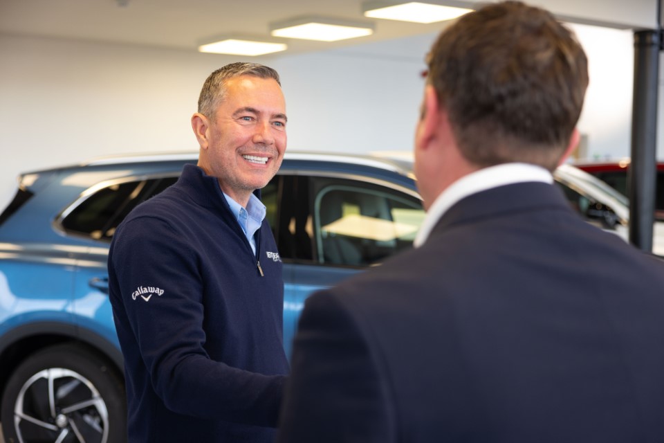 NextGear Capital employee wearing a dark blue jumper shaking hands with a customer with a light blue car in the background.