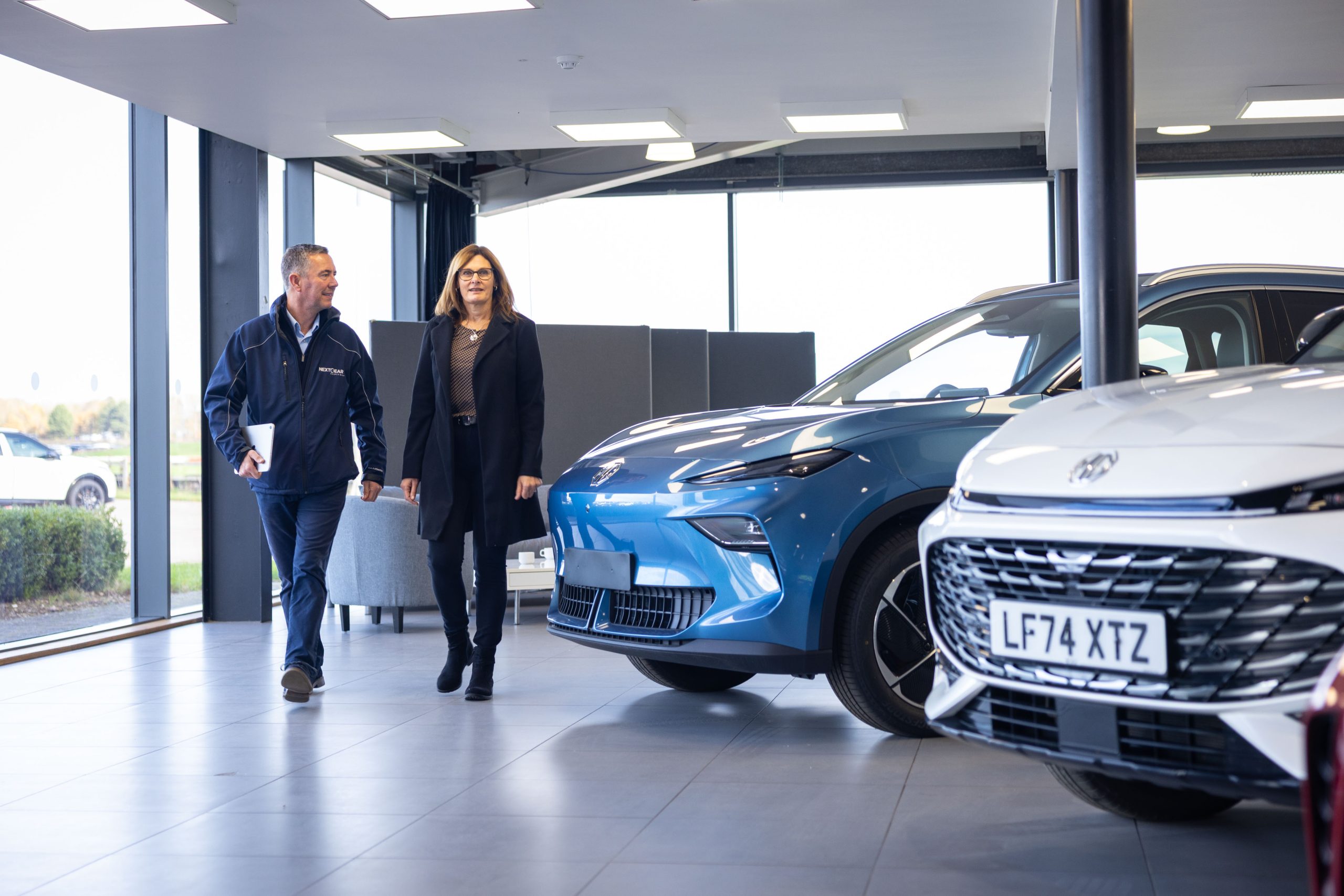 Two people walking through a car showroom with a blue car in the back and a while car in front.